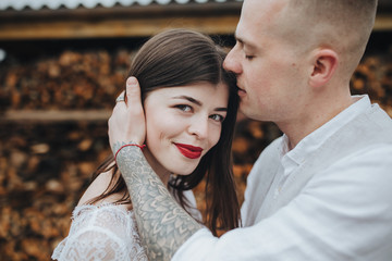 Spring wedding in the mountains. A guy in a shirt and vest and a girl in a white dress and with a bouquet in their hands are standing near a barn with firewood