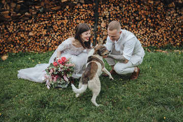 Spring wedding in the mountains. A guy in a shirt and vest and a girl in a white dress and with a bouquet in their hands are standing near a barn with firewood