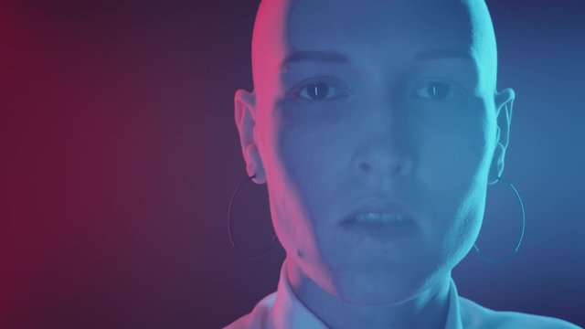 Handheld camera shot of young emotionless woman with shaved head turning face to camera and posing against dark background in studio with multicolored neon light