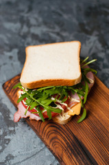 Very tasty big sandwich with ham or balyk, arugula, cheese, tomatoes and mayonnaise on a cutting board in rustic style on a gray background view from the top, copy space