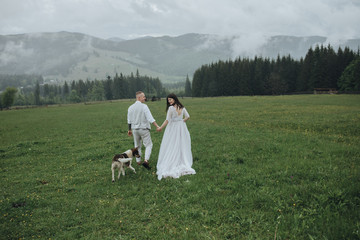 Spring wedding in the mountains. A guy in a shirt and vest and a girl in a white dress with a dog are walking along a green meadow against the backdrop of mountains and forest