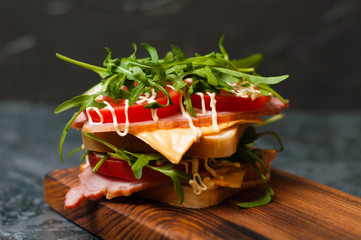 Very appetizing tasty large sandwich with ham or balyk, arugula, cheese, tomatoes and mayonnaise on a cutting board in a rustic style on a gray background view from the side, copy space