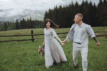 Spring wedding in the mountains. A guy in a shirt and vest and a girl in a white dress walk along a wooden fence along a green meadow against the backdrop of mountains and forests