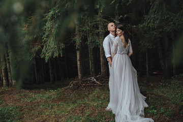 Fototapeta premium Spring wedding in the mountains. A young guy in a white shirt and trousers and a girl in a white dress are standing under the trees in a pine forest