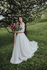 Obraz na płótnie Canvas Spring wedding in the mountains. A young girl in a white dress stands near a blossoming tree in the mountains and holds in her hands a bouquet of red flowers with a ribbon