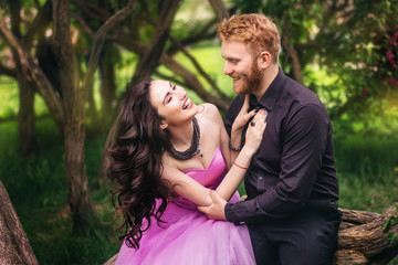 Beautiful couple in love cuddling in a blooming lilac garden. A woman in a purple sleeveless dress with a delicate necklace around her neck. Red-haired man in a black suit kisses and hugs a girl