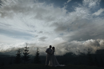 Spring wedding in the mountains. A young guy in a white shirt and trousers and a girl in a white dress stand on a mountain peak against the background of sky and trees