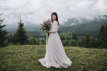 Fototapeta premium Spring wedding in the mountains. A young girl in a white dress stands in a green meadow in the mountains and holds in her hands a bouquet of red flowers and greenery