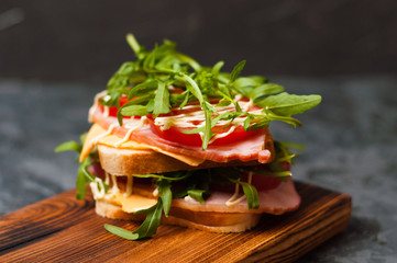 Very appetizing tasty large sandwich with ham or balyk, arugula, cheese, tomatoes and mayonnaise on a cutting board in a rustic style on a gray background view from the side, copy space