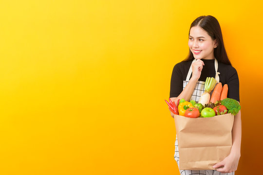  Beautiful young woman is holding vegetables in grocery bag in studio yellow  background