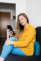 Beautiful adult female sitting on sofa while using smartphone