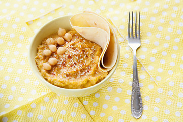 Yellow bowl of hummus, fork on yellow background. Homemade traditional hummus with paprika, olive oil, sesame seed, piece of bread. Horizontal top view. Vegetarian dish.