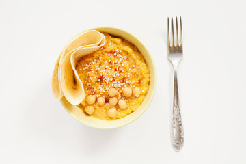 Yellow bowl of hummus, fork on white background. Homemade traditional hummus with paprika, olive oil, sesame seed, piece of bread. Horizontal top view. Vegetarian dish.