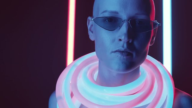 Chest up studio shot of young bald woman in trendy eyeglasses and glowing neon tubes around her neck looking at camera while posing against dark background with colorful fluorescent lamps