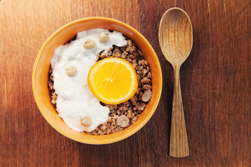 Muesli, granola with yogurt and orange in yellow bowl and wooden spoon on wooden background 