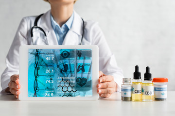 cropped view of mature doctor in white coat holding digital tablet with medical app near bottles with cbd and cannabis lettering