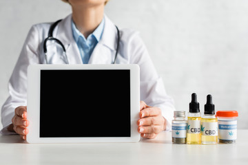 Fototapeta premium cropped view of mature doctor in white coat holding digital tablet with blank screen near bottles with cbd and medical cannabis lettering in clinic