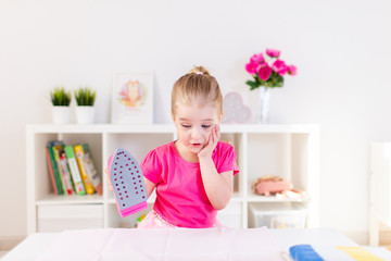 Little blonde baby girl housewife playing with toy pink iron. Ironong clothes on a white table. Housework, helper