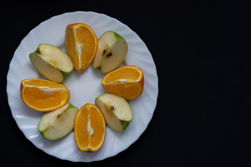 Naklejka na ściany i meble Slices of apple and orange on white plate on dark black background, top view