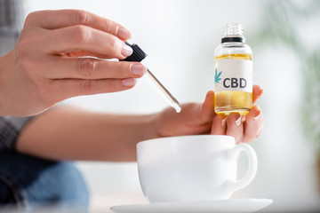 cropped view of mature woman adding drop of cbd in cup of tea on table