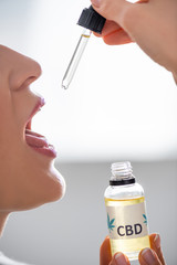 cropped view of mature woman with opened mouth holding pipette and bottle with cbd lettering