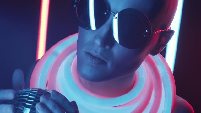 Studio shot of female fashion model in stylish glasses and glowing neon tubes around her neck holding disco ball while posing in dark room with fluorescent lamps