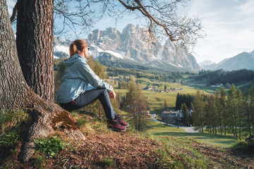 Beautiful young woman in sunglasses and blue jacket is sitting on the hill under the tree at...