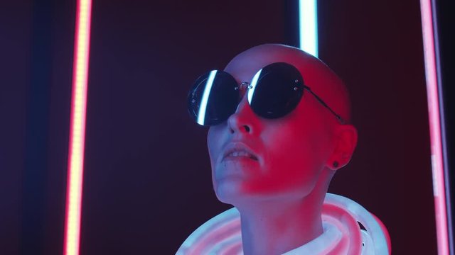 Chest up shot of bald fashion model in stylish round sunglasses and glowing neon tube looking from side to side while posing in dark studio with multicolored fluorescent lamps in background