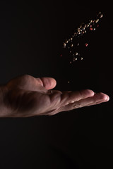 A man's hand scattering and tossing up a mix of different kinds of pepper grains on black background with strong light with his palm