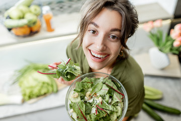 Portrait of a young and cheerful woman eating salad standing on the kitchen with food ingredients...
