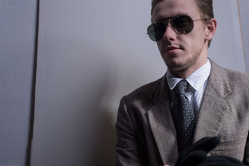 Studio portrait of a young guy, twenty-five years old, in a business stylish jacket, in sunglasses, holding black gloves in his hand. Looking to the side, on a gray background.