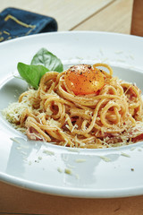 An appetizing portion of homemade Italian carbonara pasta(spaghetti) with egg yolk, bacon and parmesan in a white plate on a wooden background. Restaurant table setting