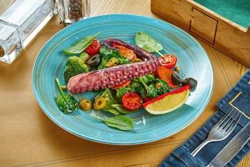 Serving of appetizing salad with octopus tentacle and steamed vegetables on a blue plate on a wooden background. Restaurant table setting
