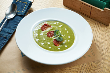 Tasty and diet cream soup with spinach and broccoli with spicy chorizo salami in a white plate on a wooden background. Restaurant table setting