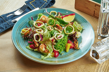 Fresh and green salad with seafood (shrimp, squid, mussels, scallops), cherry tomatoes in a blue plate on a wooden background. Restaurant table setting