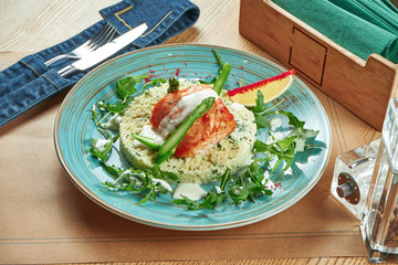 A large salmon steak with a side dish of corn polenta, fresh asparagus and arugula on a ceramic plate on a wooden background. Restaurant table setting
