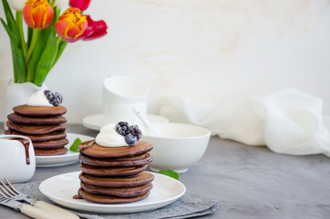 Stack of chocolate pancakes on a white plate with chocolate sauce, whipped cream, blueberries and blackberries on top of a concrete background. Delicious breakfast. Horizontal orientation. Copy space.