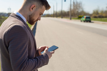 Portrait of a young bearded guy of twenty-five years old, using a smartphone, stands at the carriageway, against the background of a car