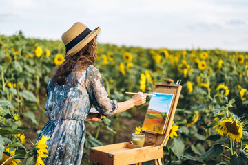 A young woman with curly hair and wearing a hat is painting in nature. A woman stands in a...