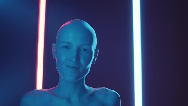 Chest up shot of young shirtless woman with hairless head turning face to camera and smiling while posing in dark studio with neon light