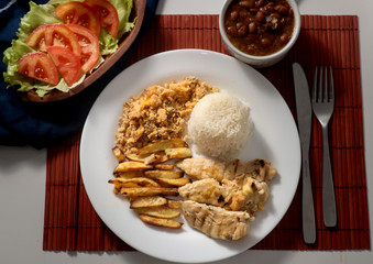 Dish of grilled chicken fillet with rice, beans, french fries and farofa on white plate. Top view.