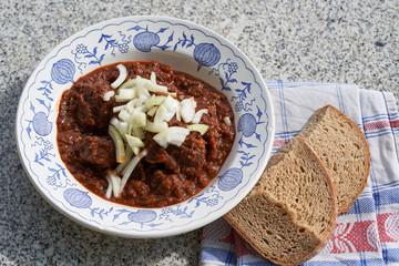 Traditional hungarian style beef goulash served with chopped white onion on the top in rustic porcelain deep plate and two slices of dark bread. Tasty and spicy dish from east european countries.