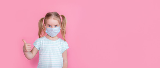 Little blonde girl with two ponytales in a medical mask face on a pink background showing thumbs up. Copyspace.