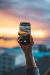 Hand holding a smartphone taking a photograph of the sea at sunset - obrazy, fototapety, plakaty