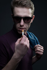 close up portrait of a young guy wearing sunglasses looking at the camera while holding a cigarette in his hand, preparing to light a cigarette. Denim style. on a gray background.