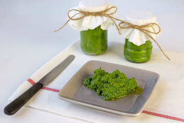 Homemade fresh pesto sauce on a plate with glass jars aside on white cloth on the kitchen table, with knife  aside. Fresh, healthy food. Homemade preserves.
