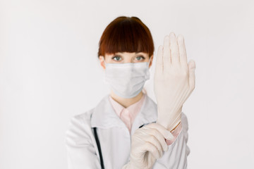 Confident young woman doctor or nurse in white coat and with medical mask posing to camera and showing how to protect hands with rubber gloves. Isolated on white, copy space, focus on hand