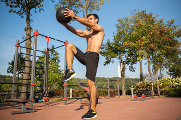 Fit guy doing exercises using a ball outdoors. Young athletic man training in city park