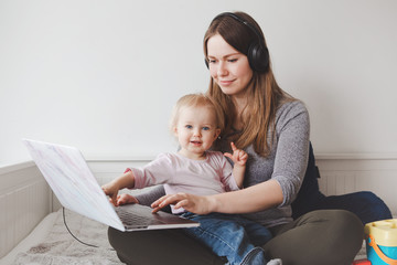 Young Caucasian mother with toddler baby working on laptop from home. Workplace of freelance woman...