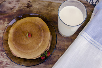 Top view of pile of pancakes with strawberries and glass of fresh milk on village wooden table with linen napkin. Concept of countryside breakfast, summer breakfast with berries from garden.